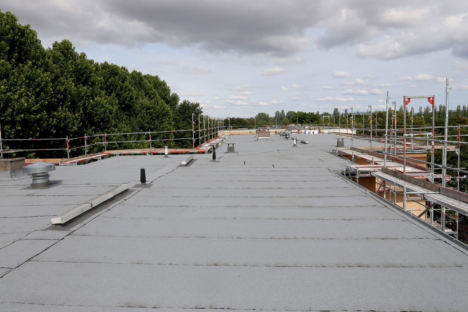 Flat, gray rooftop with various vents and pipes, surrounded by trees and a cloudy sky.