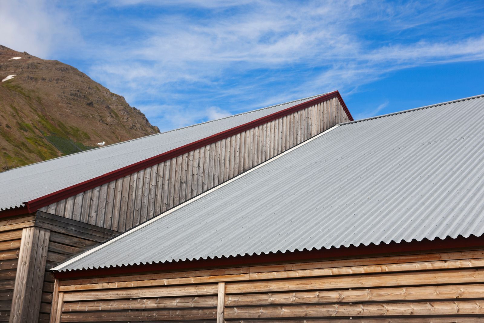Wooden buildings with corrugated metal roofs against a mountain and blue sky.