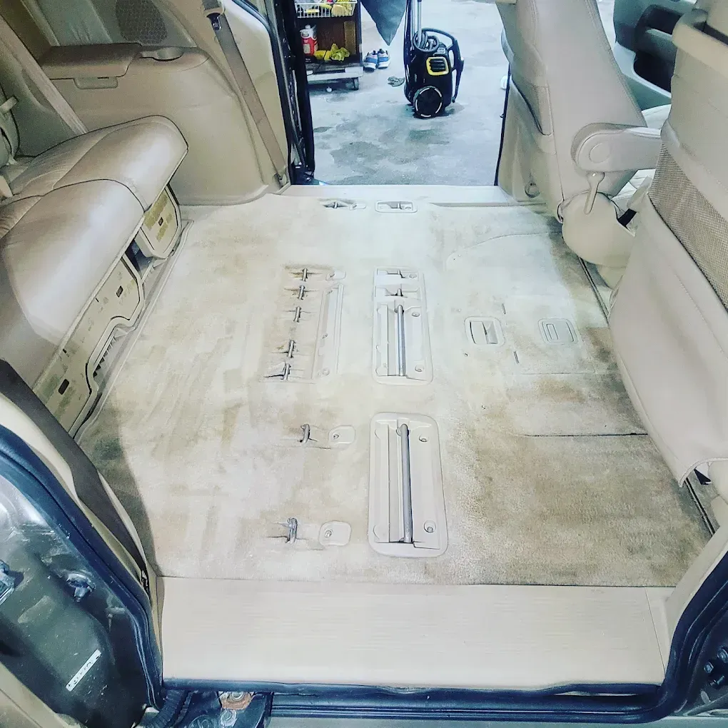 An interior view of a vehicle with seats removed, showing beige carpet, seat mounting tracks, and an open doorway.