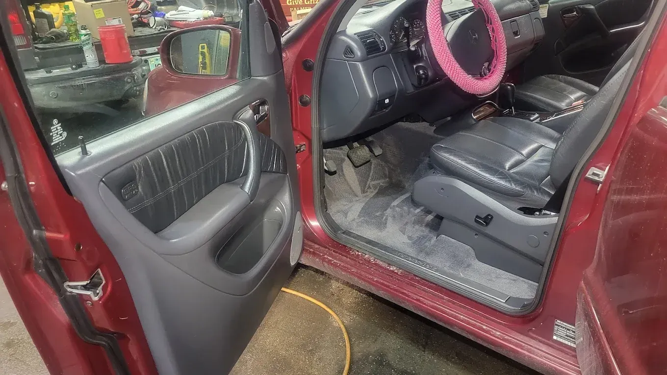 Open driver-side door of a red vehicle showing a black leather interior, gray flooring, and a pink steering wheel cover.