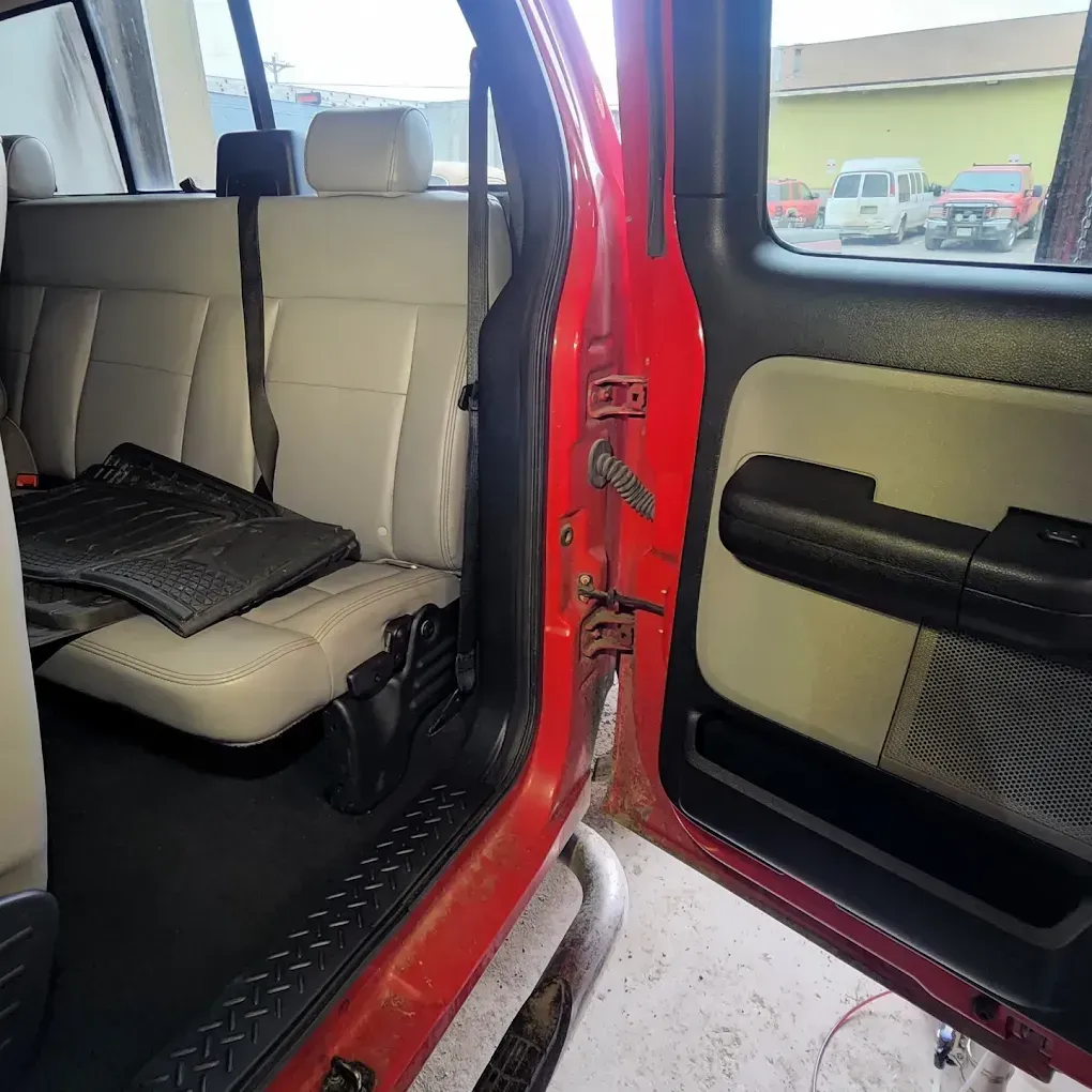 The open door of a red vehicle shows a beige interior with a seat and floor mat, viewed from inside the cabin.