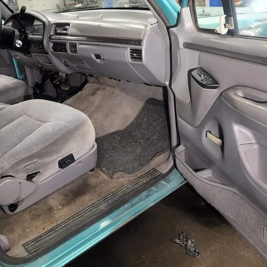 The passenger side interior of a turquoise truck featuring grey fabric seats and a grey dashboard with a worn floor mat.