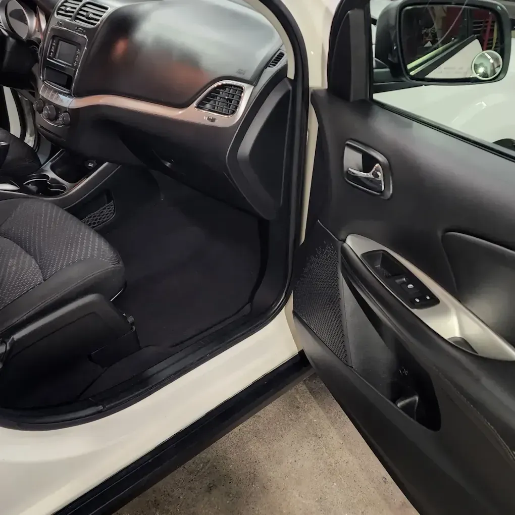 The interior of a white car, showing the open passenger door, black dashboard, fabric seat, and side mirror.