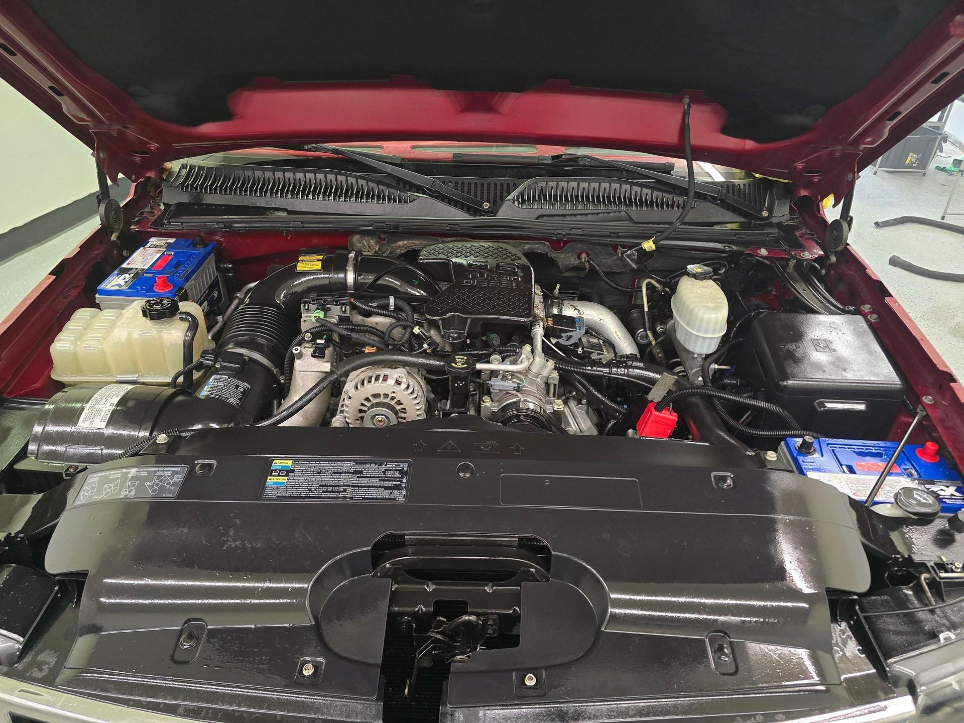 An open hood revealing a clean, dark engine bay of a maroon vehicle with two blue batteries on either side.