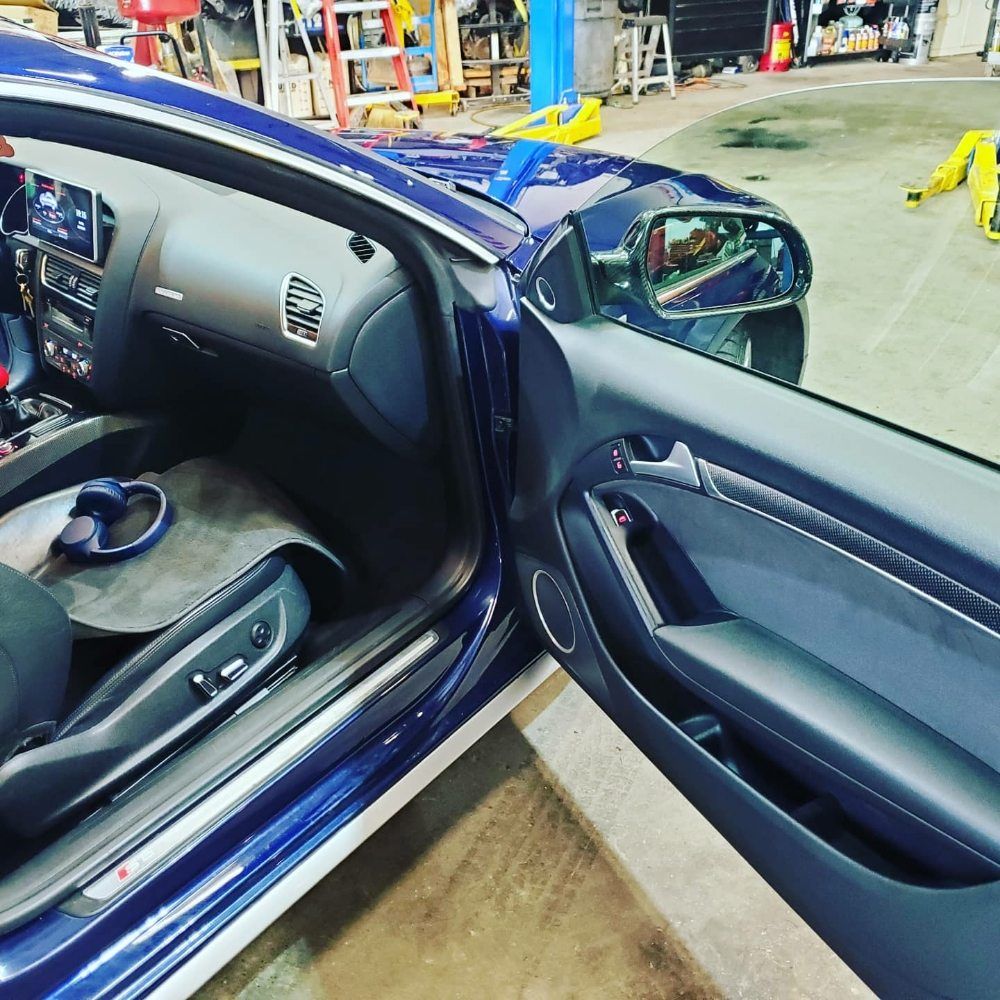 Open car door showing dark blue sedan interior, dashboard, and side mirror in a workshop.