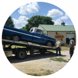Blue vintage truck being loaded onto a tow truck, near a Sunoco station. Two people nearby.