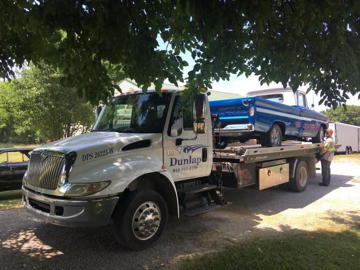Tow truck with a blue classic pickup truck on its flatbed, parked on a paved area, under trees.