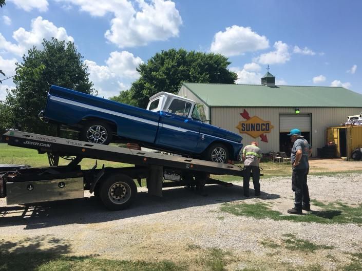 Blue and white pickup truck being unloaded from a tow truck, two men standing nearby, building in the background.