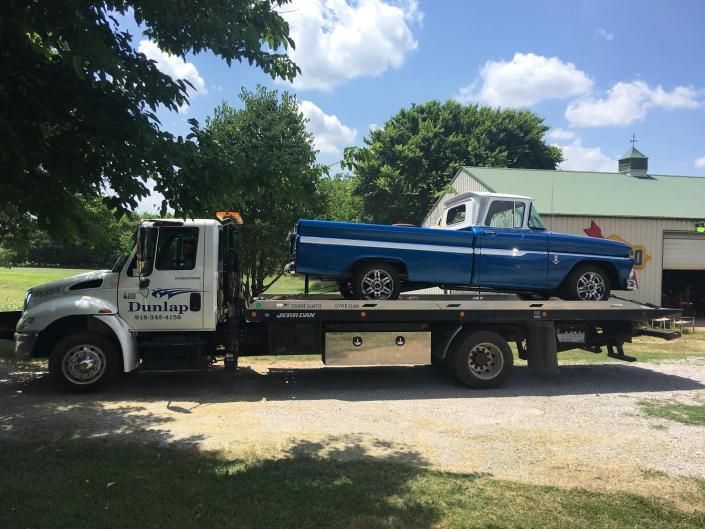 Blue vintage pickup truck on a tow truck; sunny outdoor setting, green foliage.