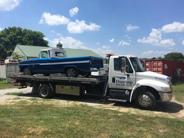 Blue classic pickup truck on a white tow truck, parked on grass, under a blue sky.