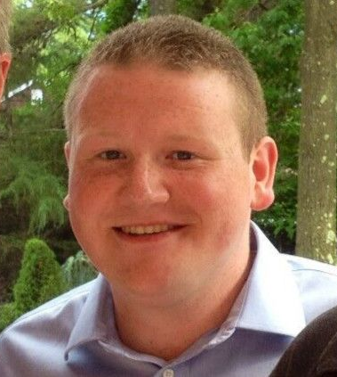 Man in a blue button-down shirt smiles, posing outdoors with trees in the background.