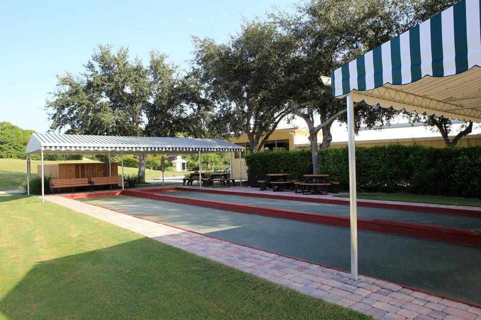 Two outdoor shuffleboard courts under striped awnings, next to a grassy lawn and benches on a sunny day.