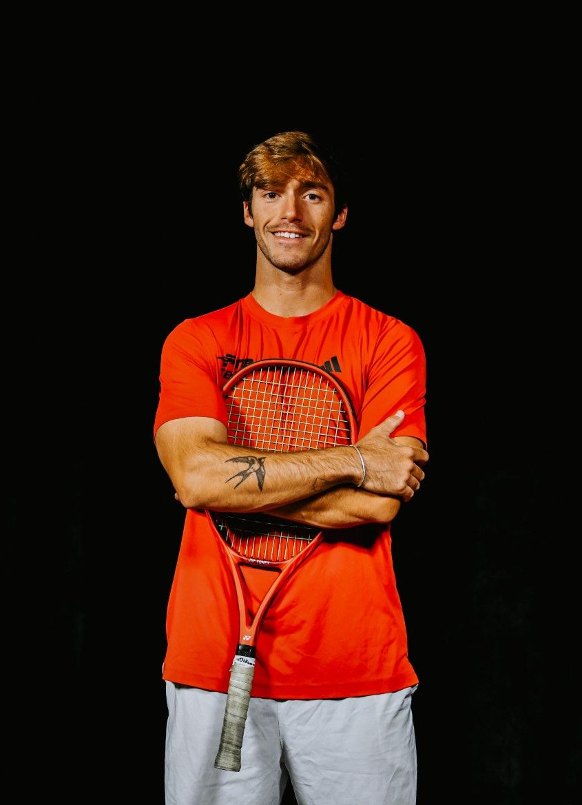A person in an orange t-shirt holding a tennis racket with crossed arms against a black background.