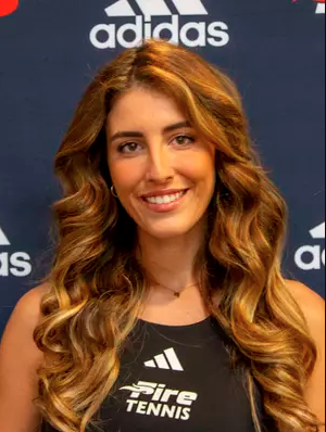 A smiling woman with long, wavy brown hair wears a black athletic tank top in front of an Adidas-branded backdrop.
