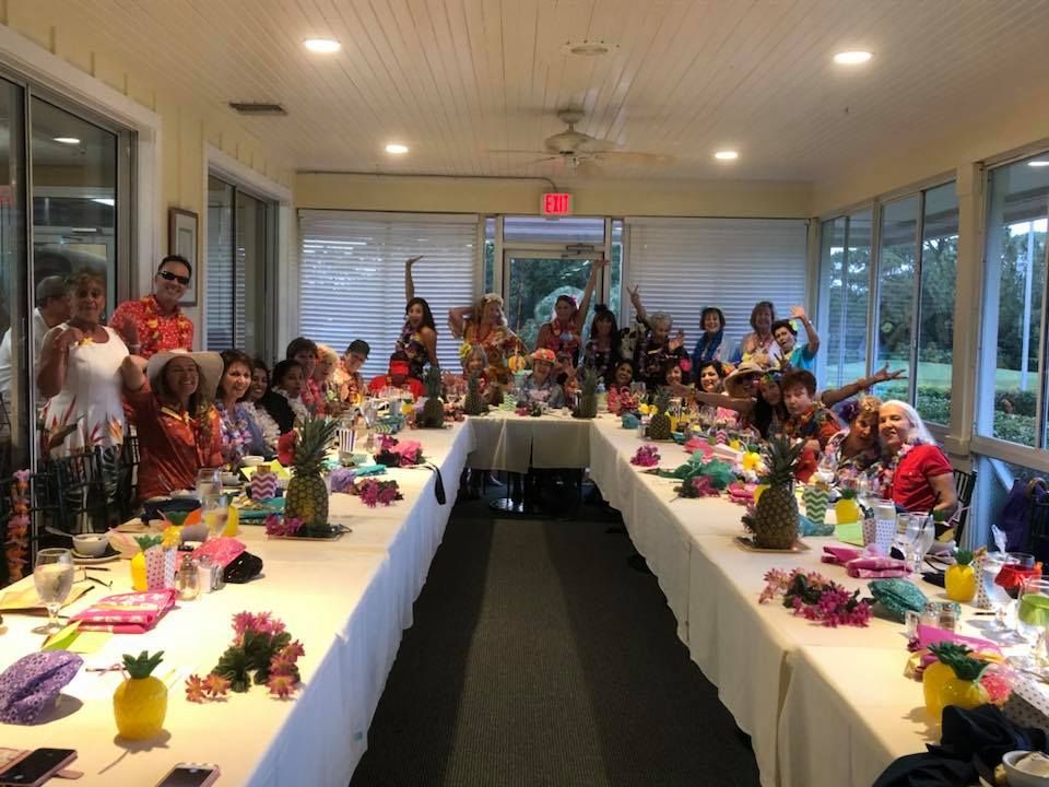 A large group of people celebrating at long tables in a sunlit dining hall decorated with pineapples and tropical items.