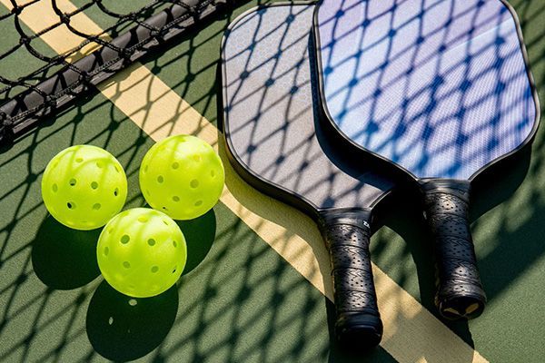 Two pickleball paddles and three bright yellow perforated pickleballs sitting on a court next to a net.