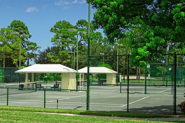 Two empty tennis courts with white shaded sitting areas and chain-link fencing, surrounded by green trees on a sunny day.