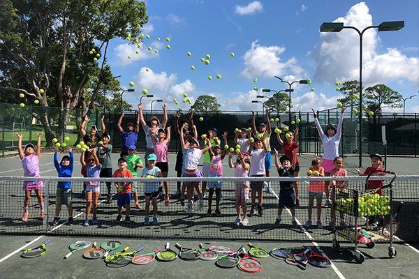 A group of children throwing tennis balls in the air on a tennis court, with tennis racquets laid out on the ground below.