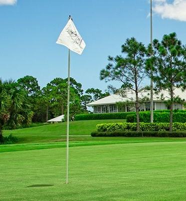 A white golf flag waves on a pole on a lush green putting green with a clubhouse and trees in the background.