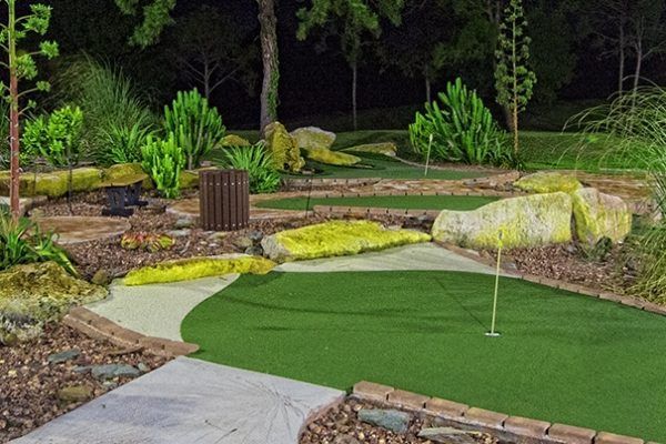 A mini-golf course at night, featuring a green putting area, large rocks, desert-style landscaping, and a flag.