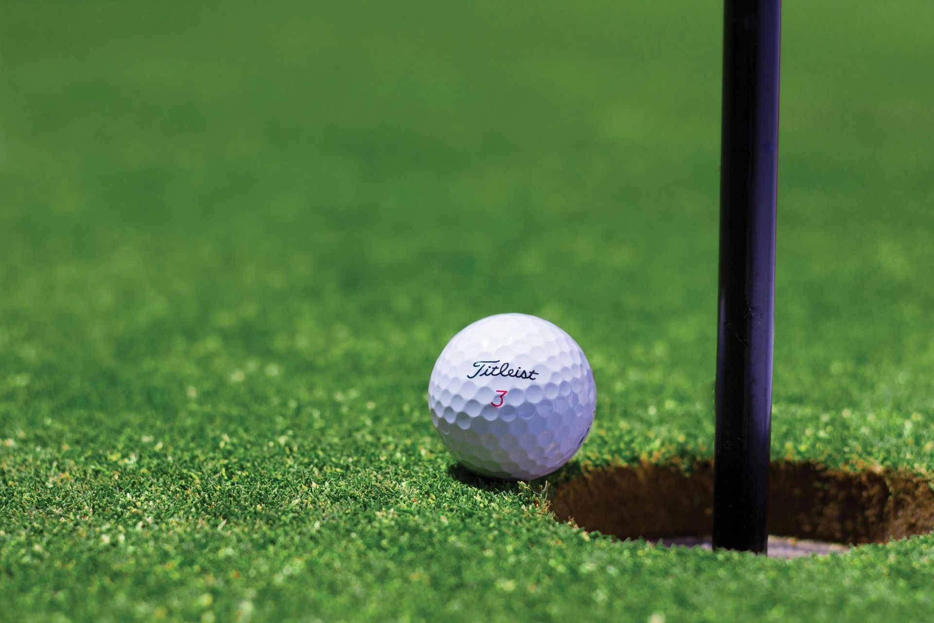 A white Titleist golf ball sits on a green grass putting surface, just inches from the edge of a hole with a flagstick.