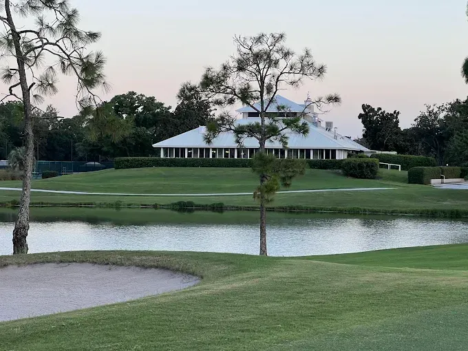 A white clubhouse sits on a grassy hill overlooking a pond and sand trap on a golf course under a pale sky.