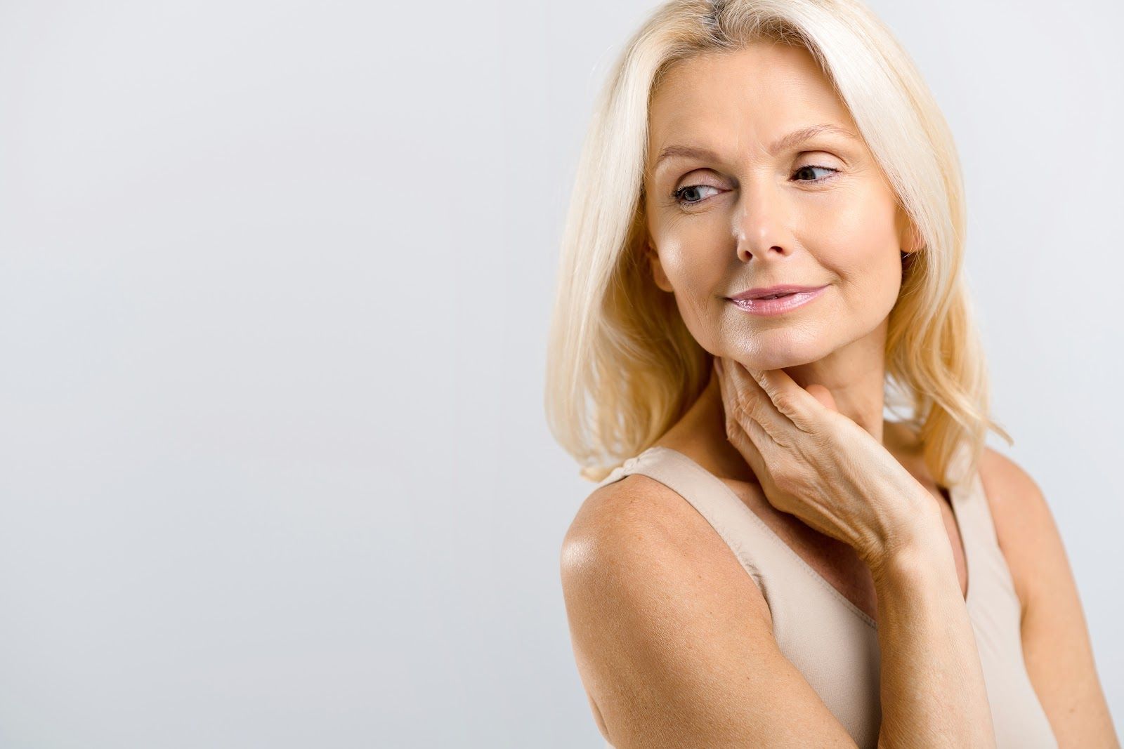 Blonde woman touches her neck, looking off to the side, wearing a beige tank top.