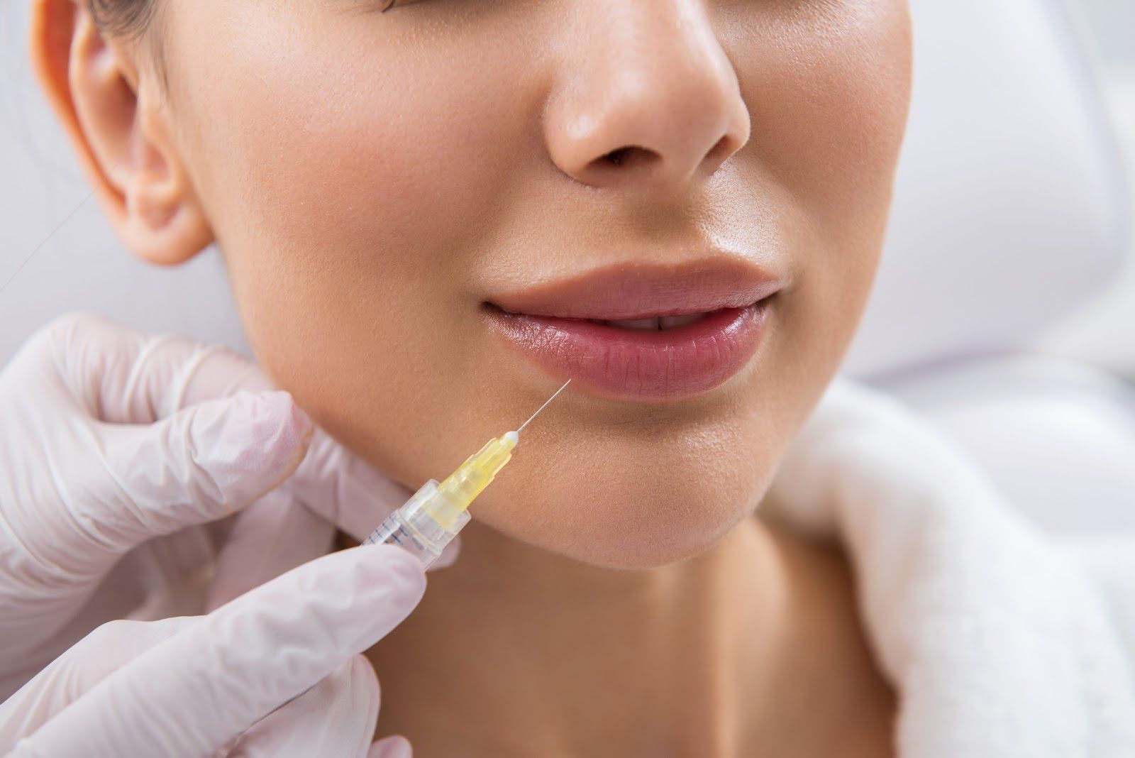 Close-up of lips receiving lip filler injection. A gloved hand holds syringe near the lip.