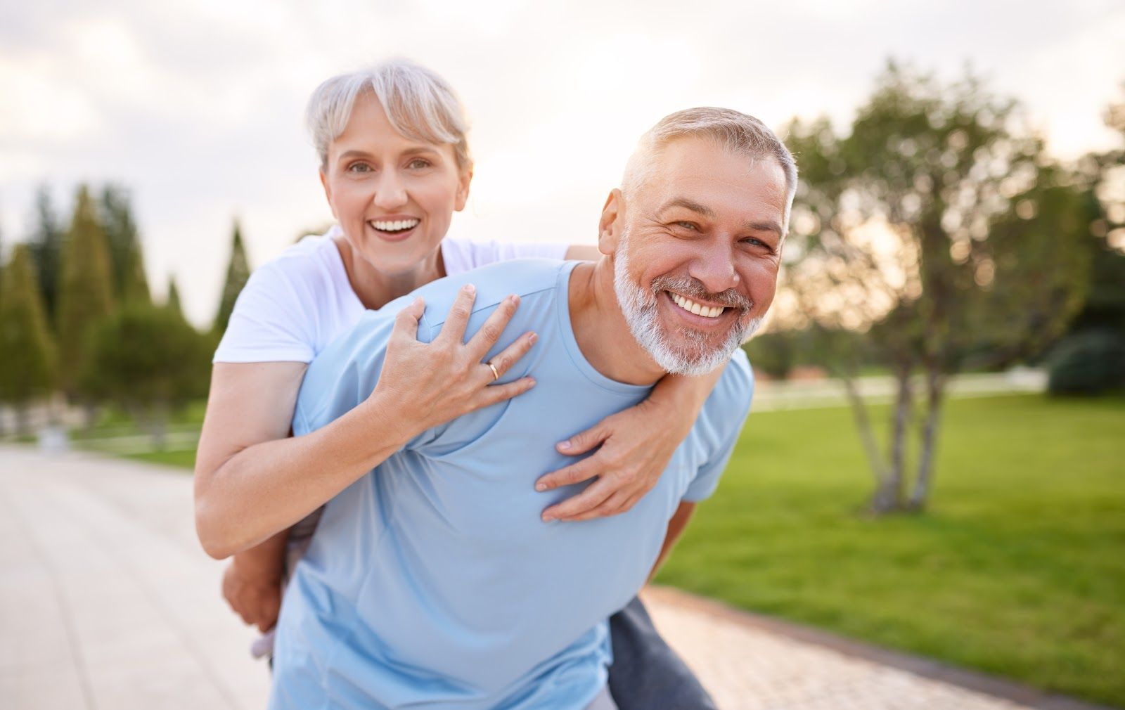 Smiling couple outdoors, woman on man’s back. Green grass, trees, and bright sky.