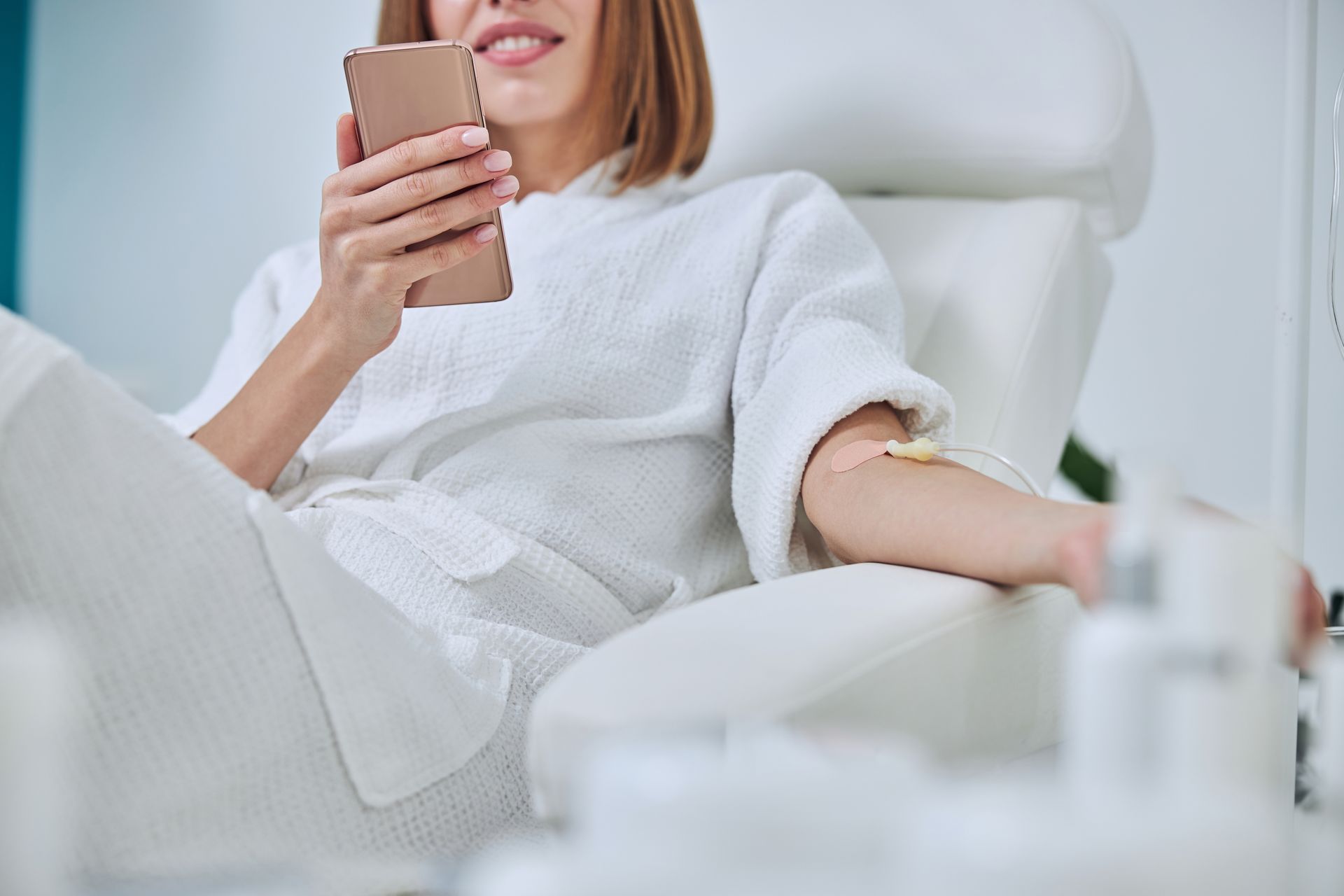 Woman in bathrobe receives IV, smiling at her phone in a spa-like setting.
