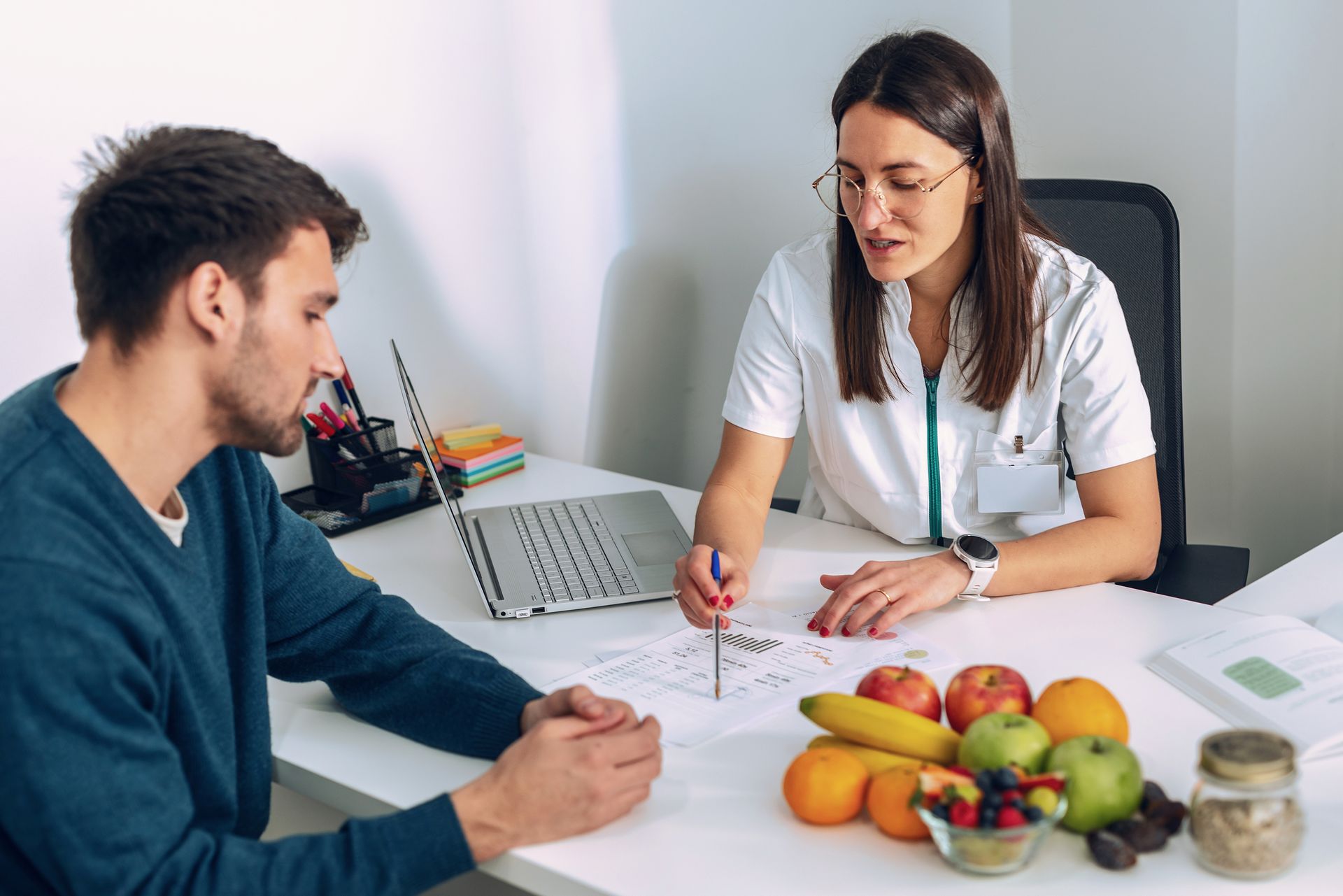 Nutritionist consults with a male patient at a desk, reviewing papers and discussing diet plans. Fruits sit on the table.