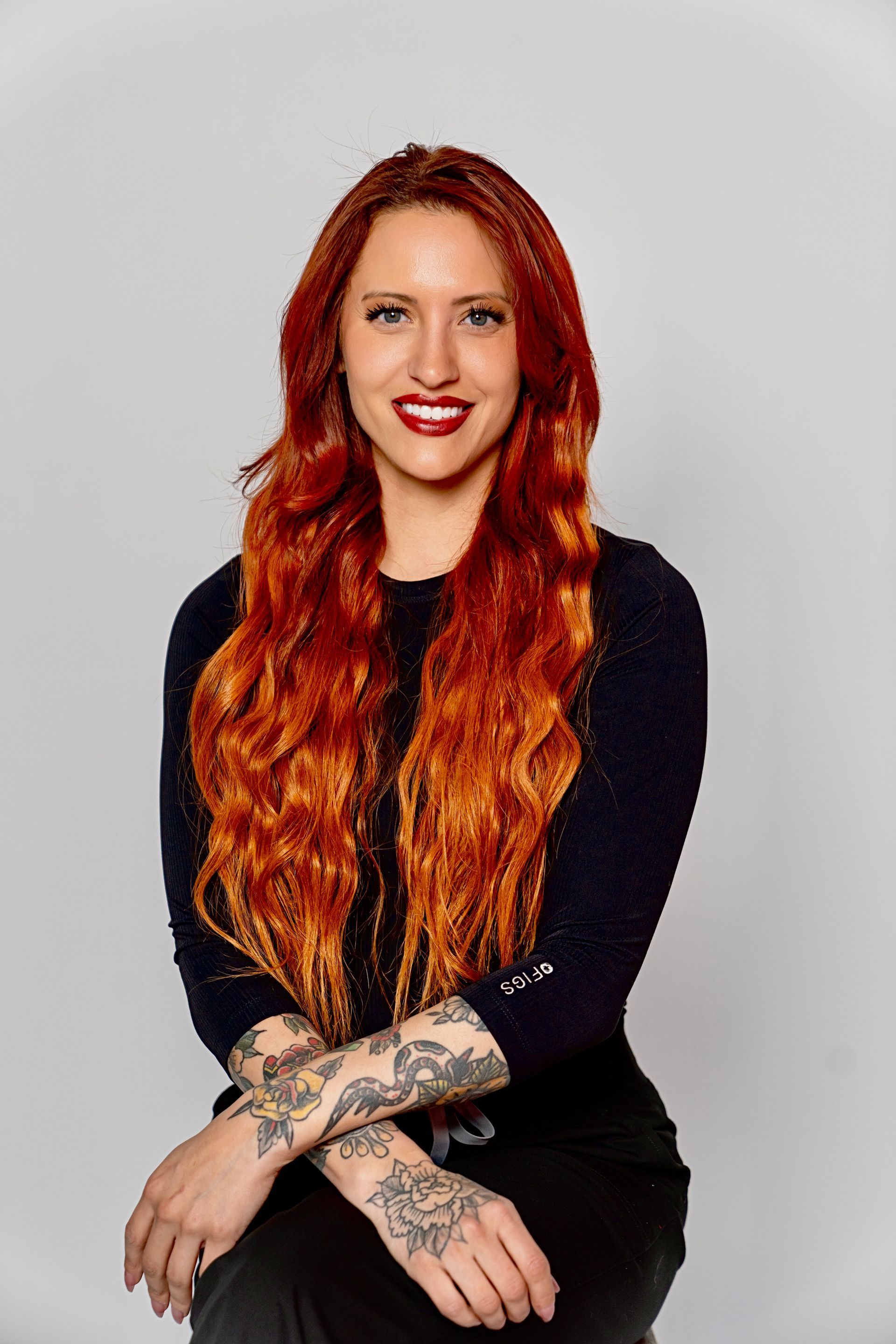 Woman with long red wavy hair, tattoos on arms, smiling, wearing dark clothing, sitting.