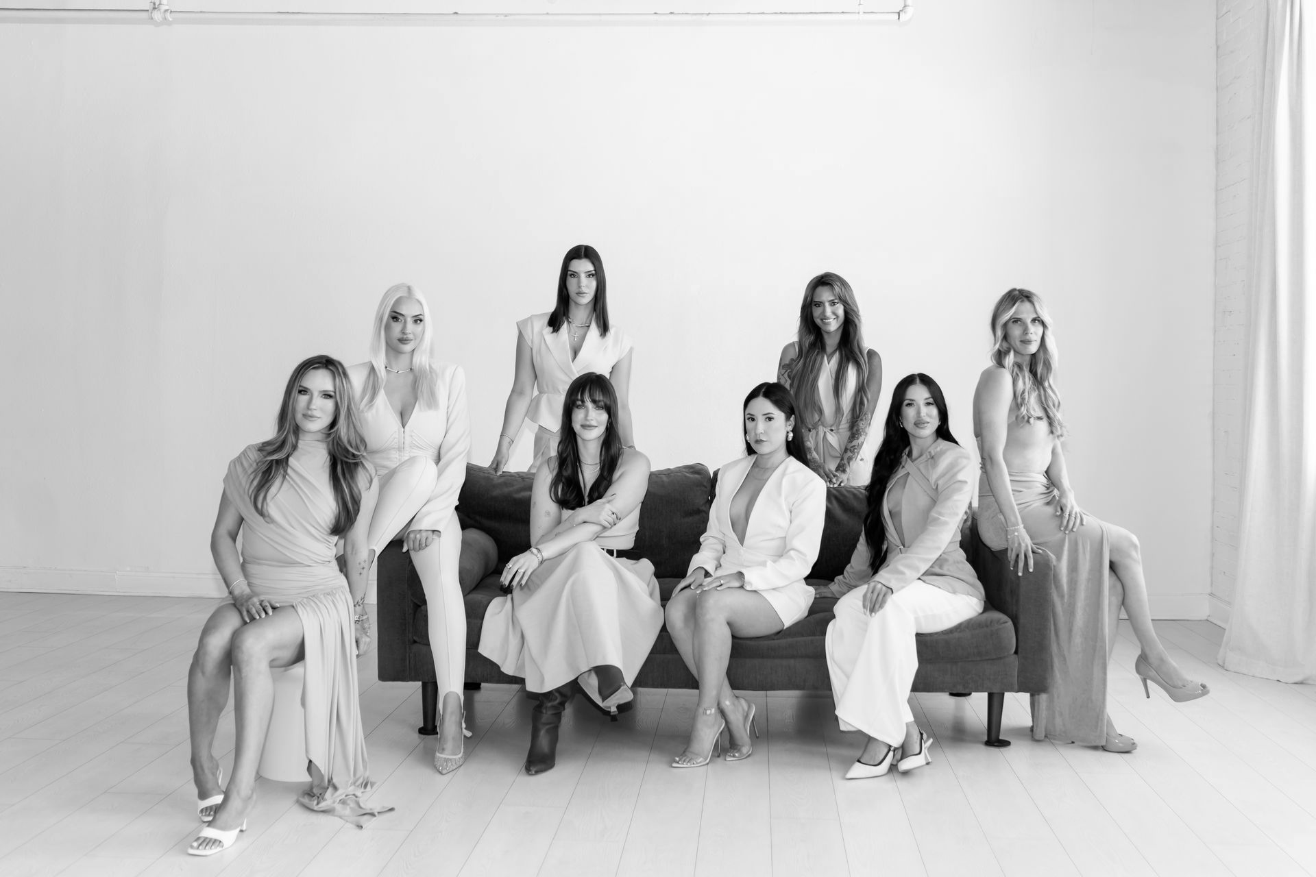 Seven women posing together on a couch in a bright studio, dressed in light-colored outfits.