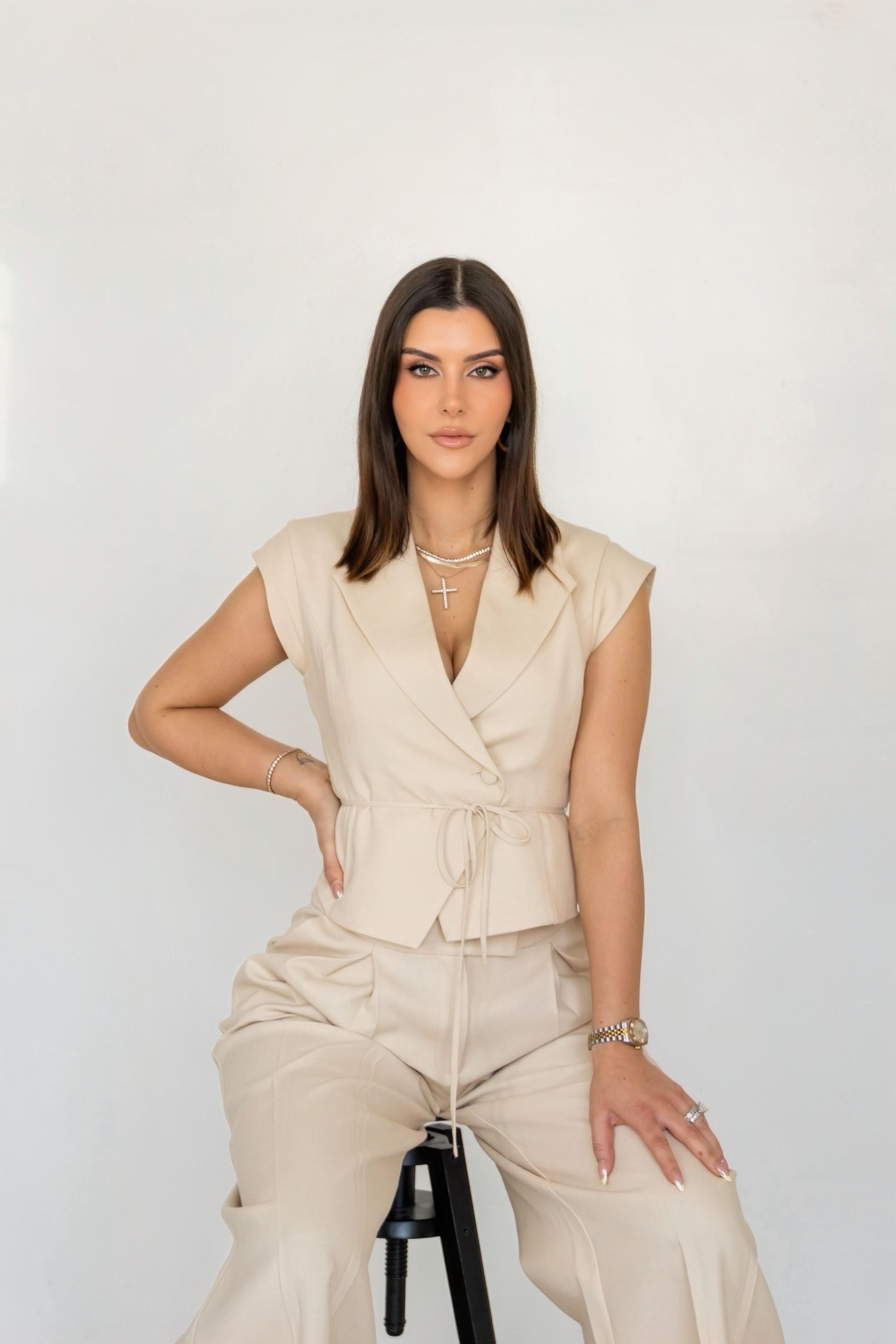 Woman seated on a stool against a white backdrop, wearing a beige blouse and matching trousers.