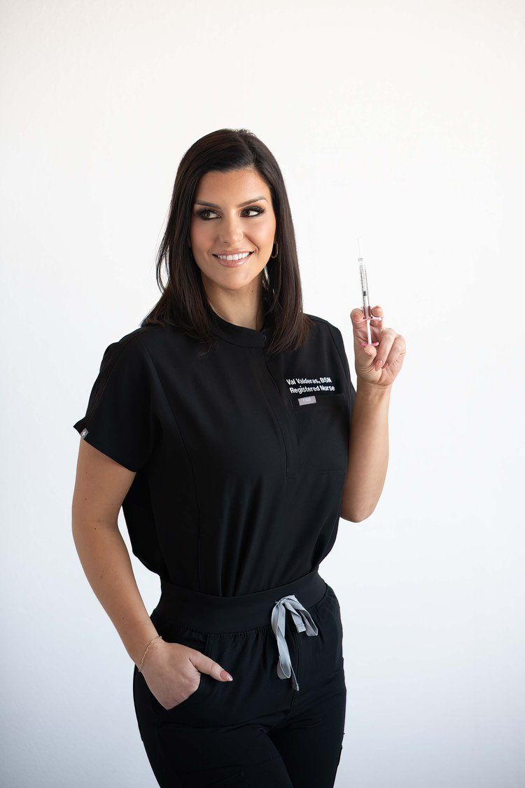 Woman in black scrubs holds a syringe, smiling, against a white backdrop.