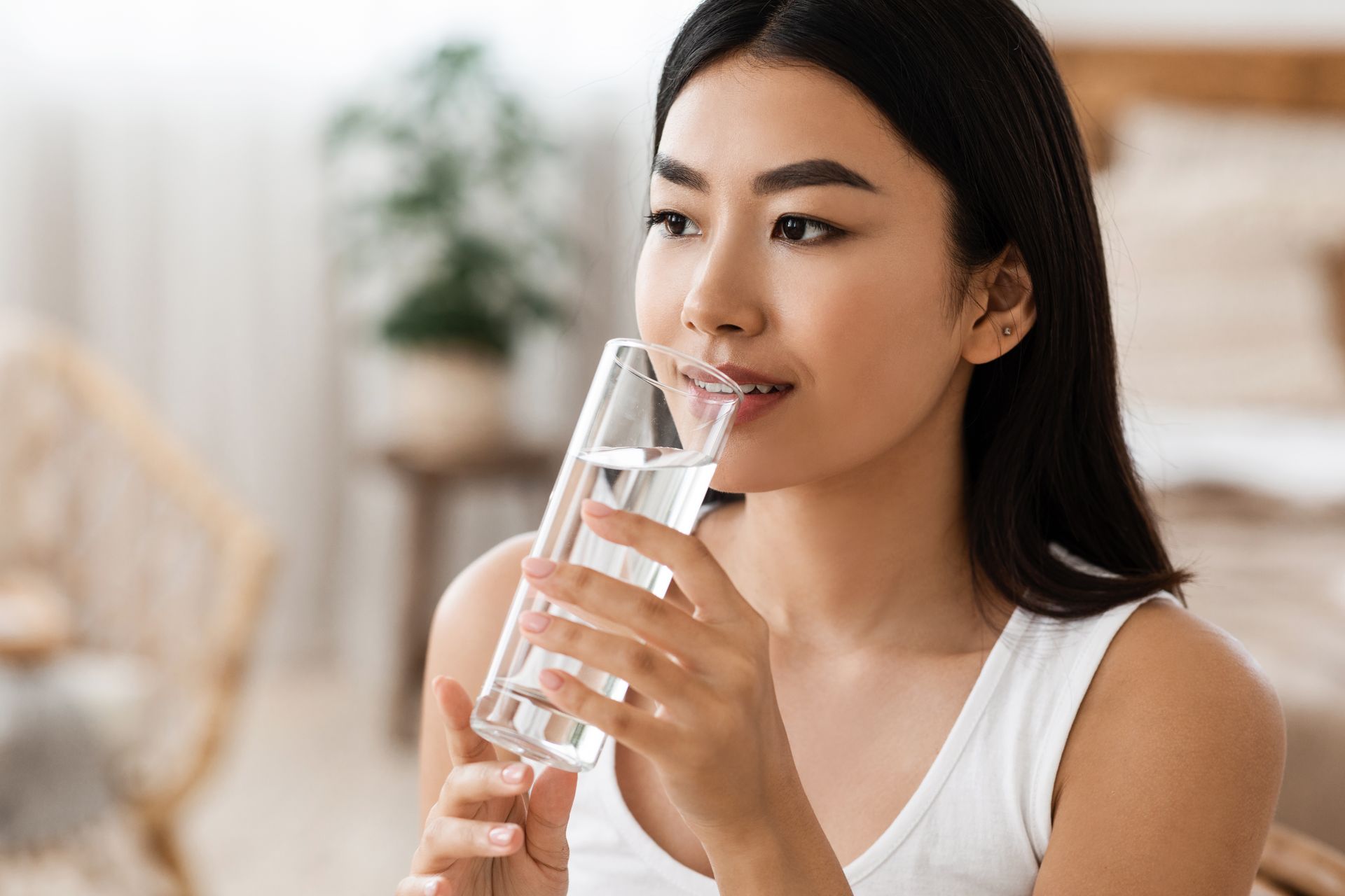 Woman drinking water from a clear glass, indoors, wearing a white tank top, looking away.