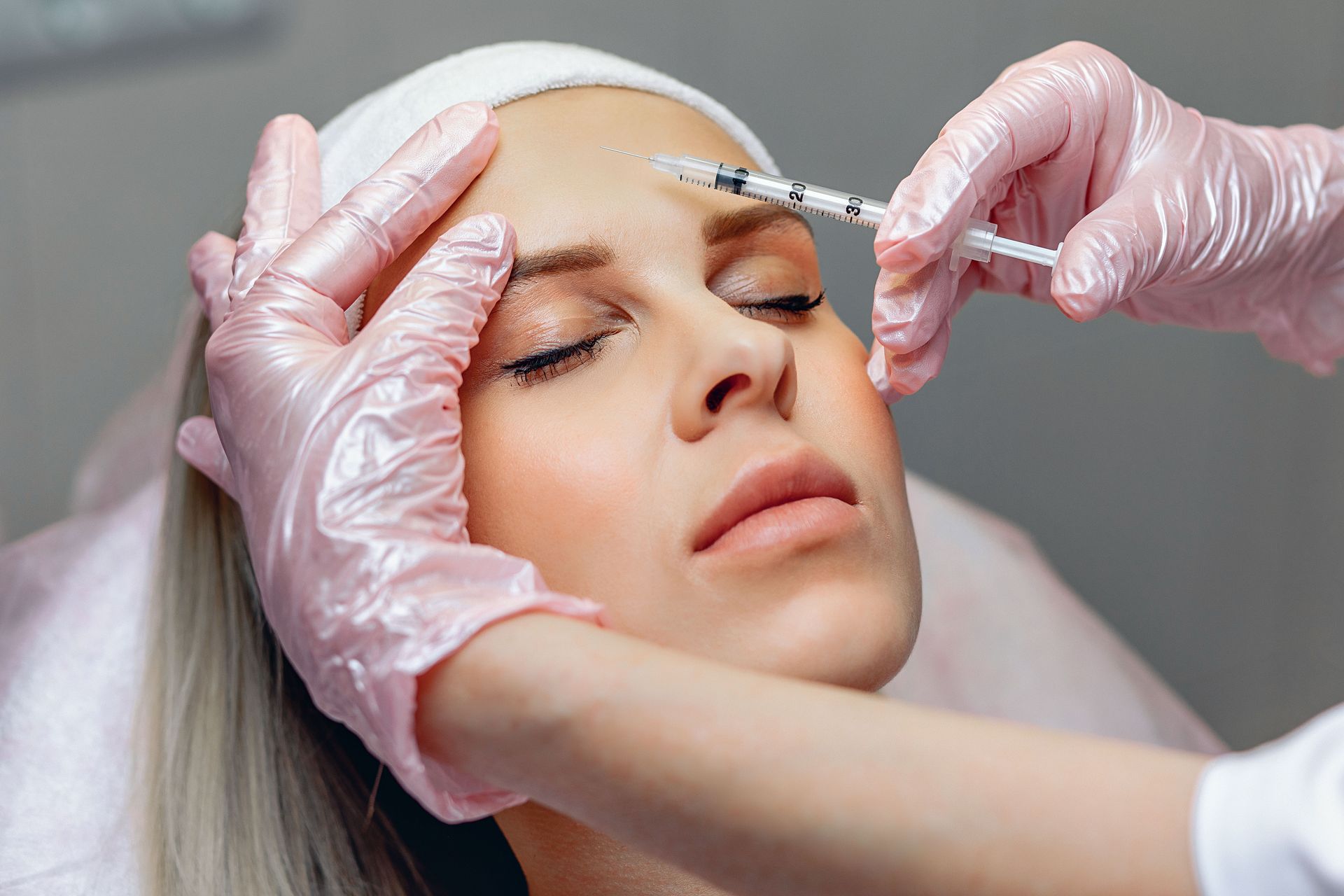 Woman receiving a forehead injection from a gloved hand; pink gloves, white headband, indoors.
