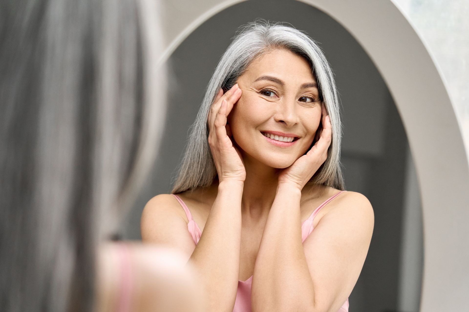 Woman with gray hair smiles in a mirror, touching her face. She wears a pink top.