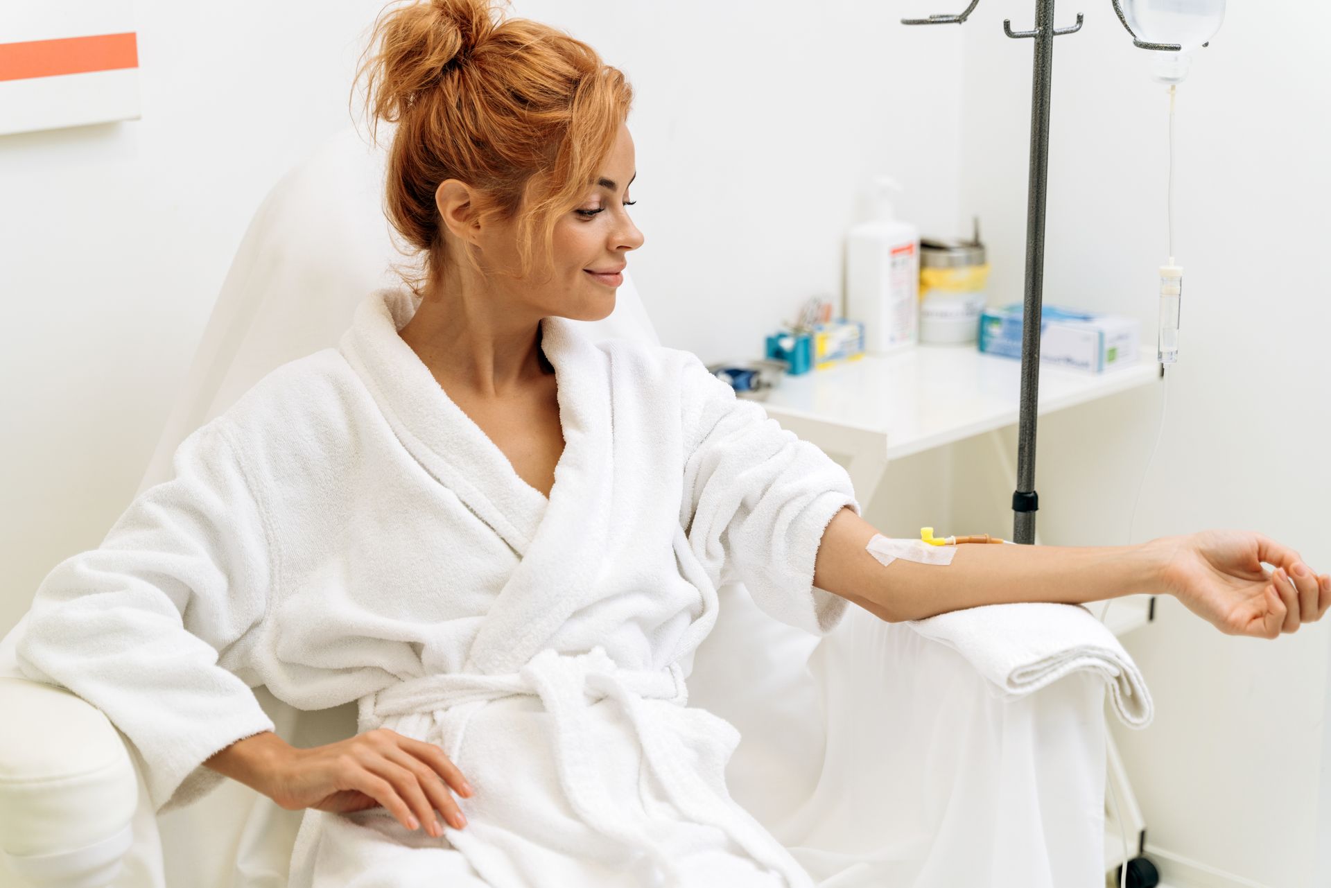 Woman in white robe receives IV drip in a medical setting, looking relaxed.