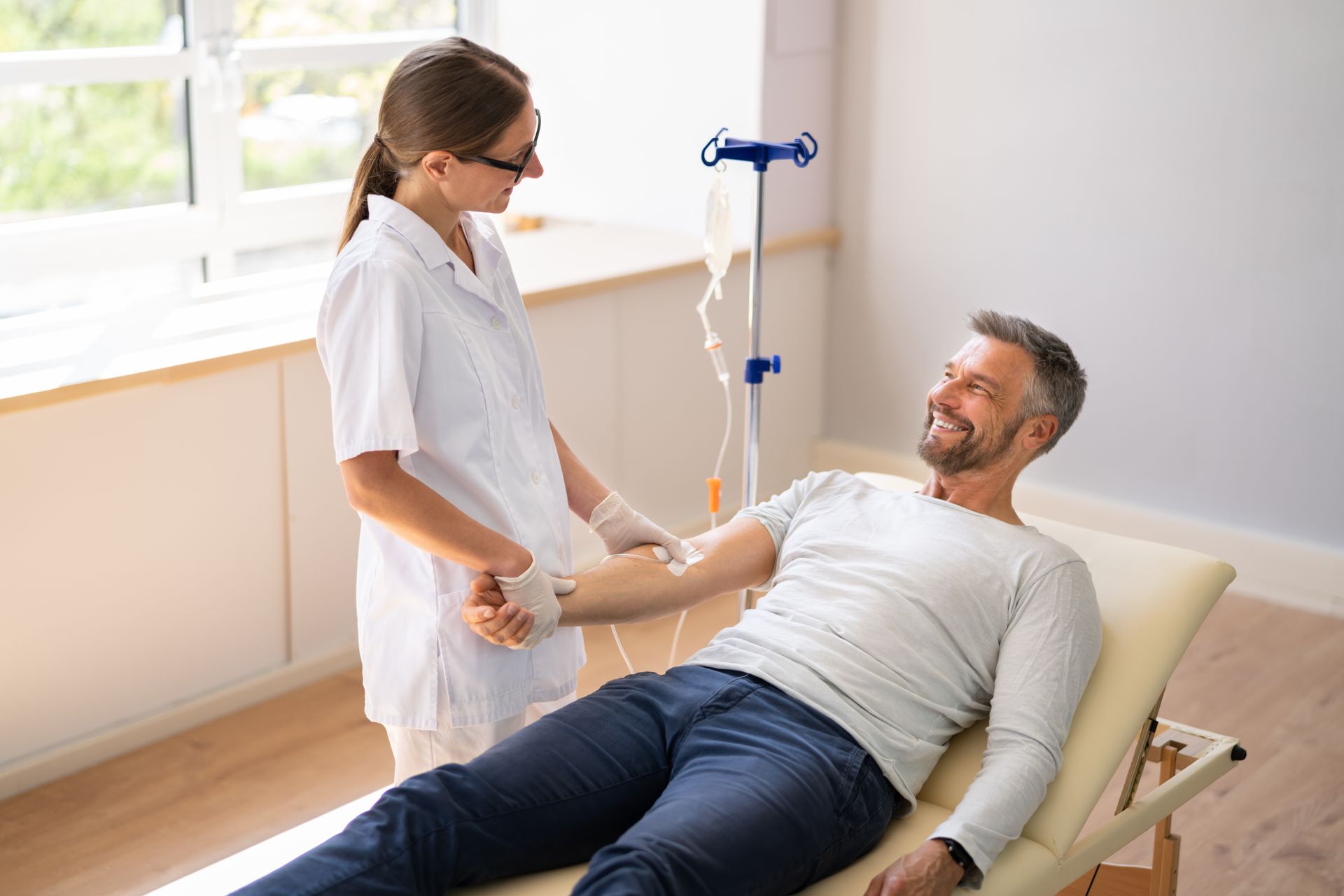Nurse giving IV fluids to a smiling man lying on a medical bed. Bright room with window.