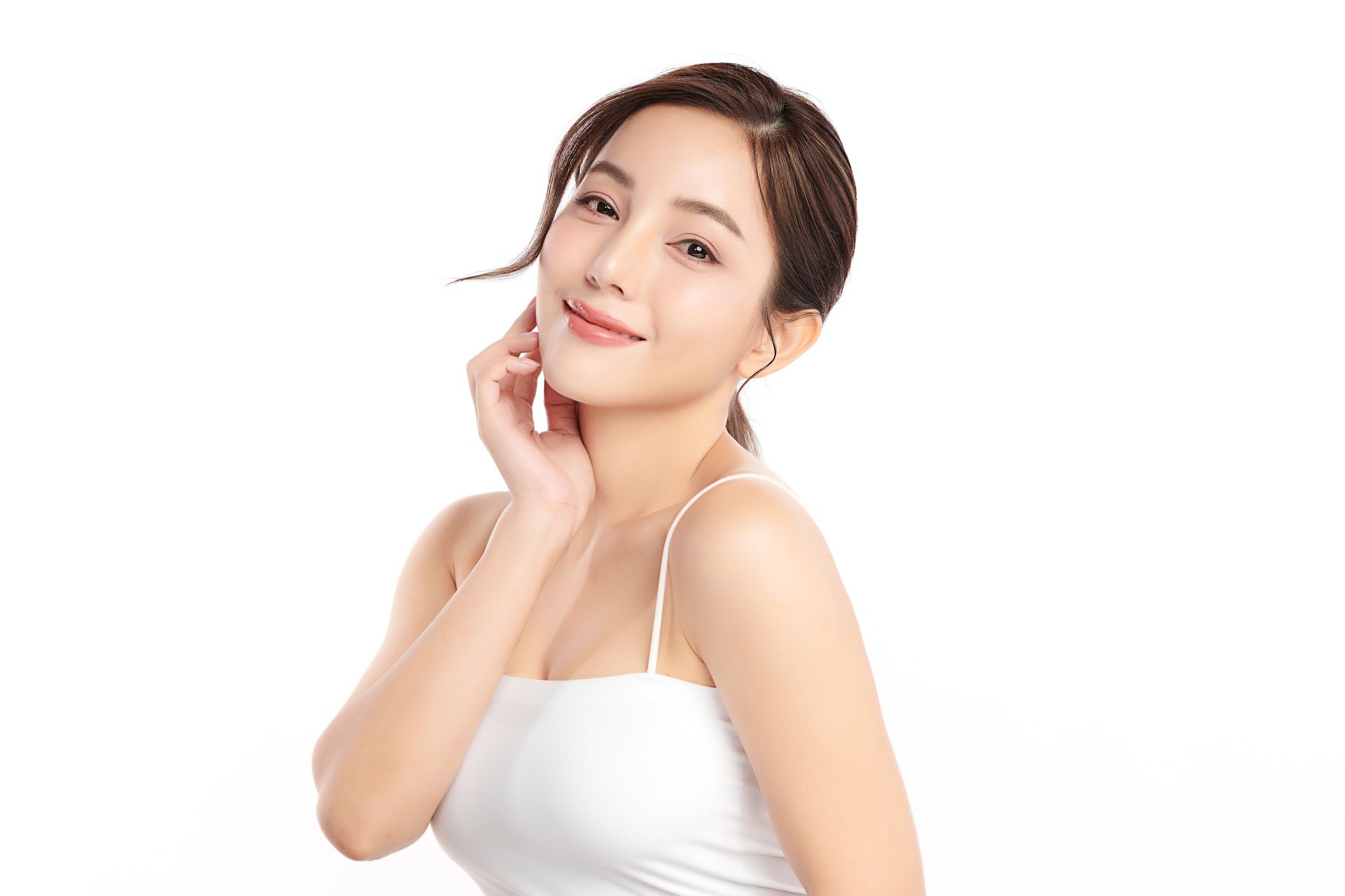 Woman in a white sleeveless top posing against a white background, smiling softly and touching her cheek.