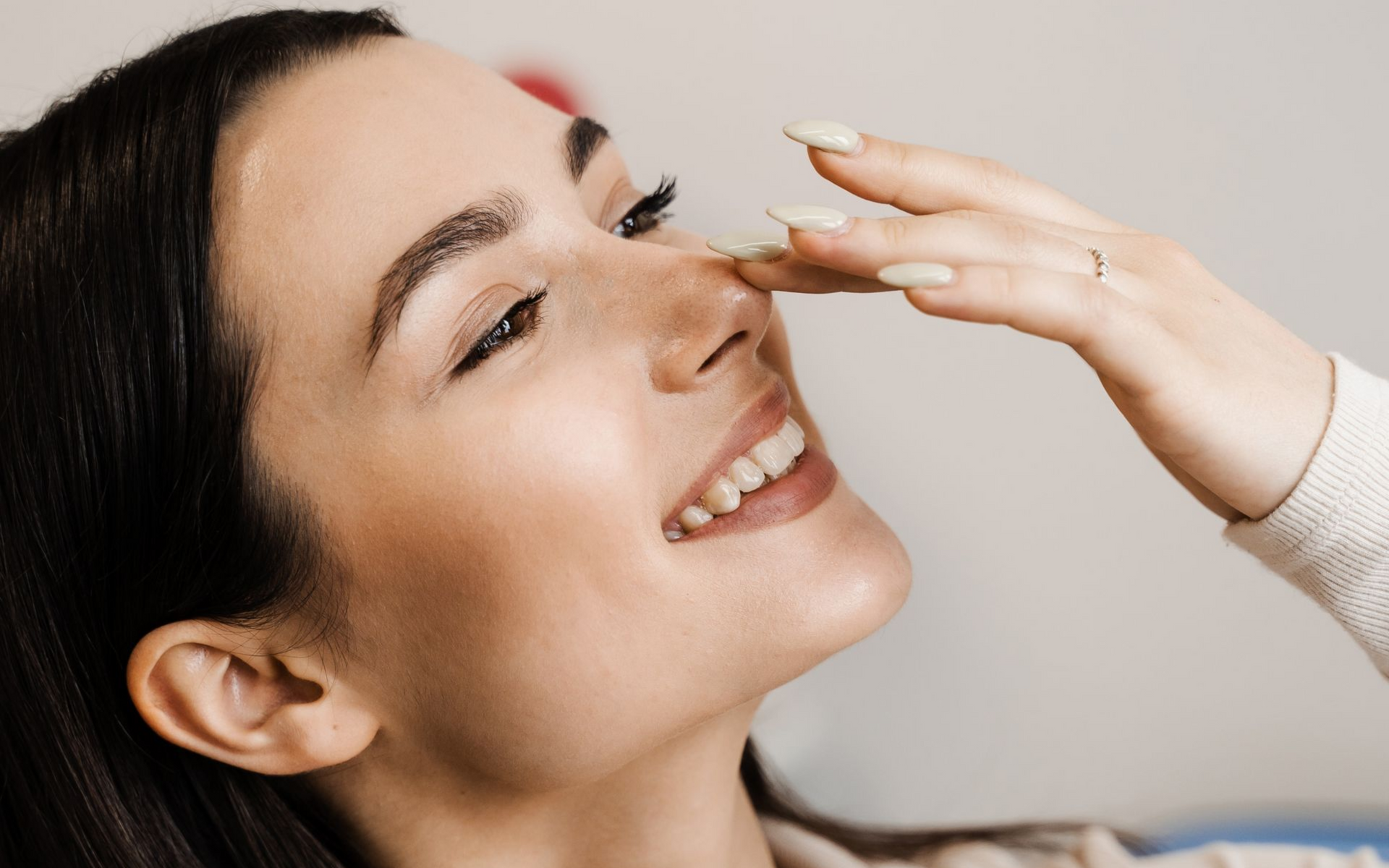 Woman smiling, touching her nose with a hand. Indoor setting, neutral background.