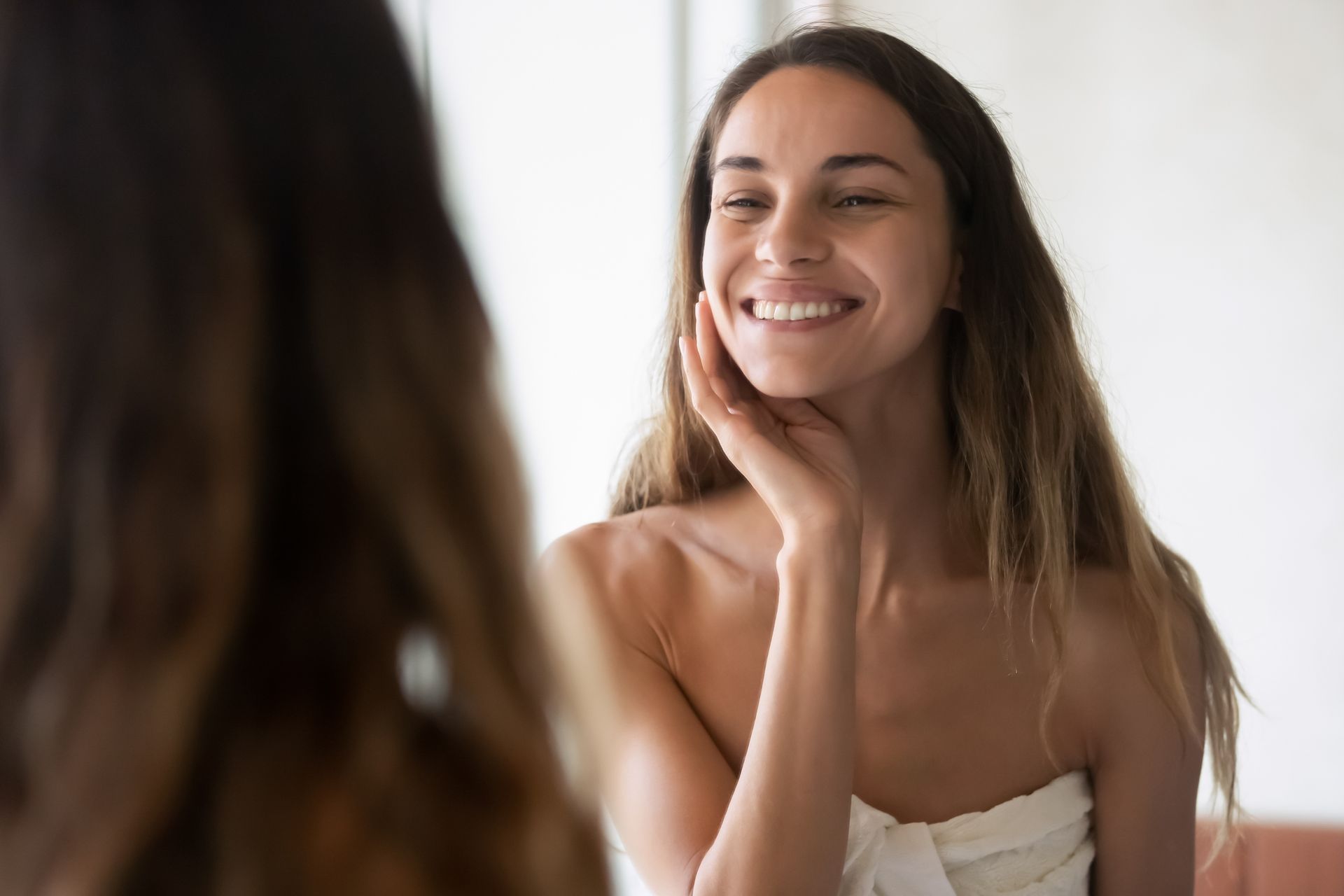 Woman smiling at herself in mirror, touching her face with a towel wrapped around her.