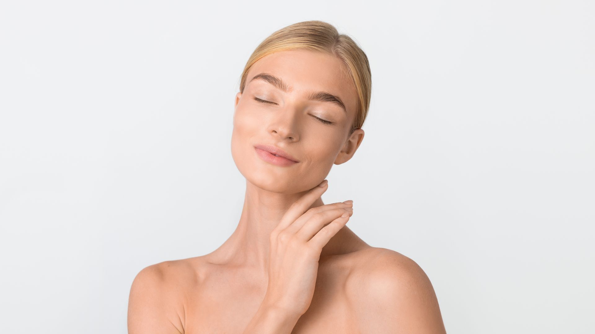 Woman with fair skin touches her neck, eyes closed, against a white background.