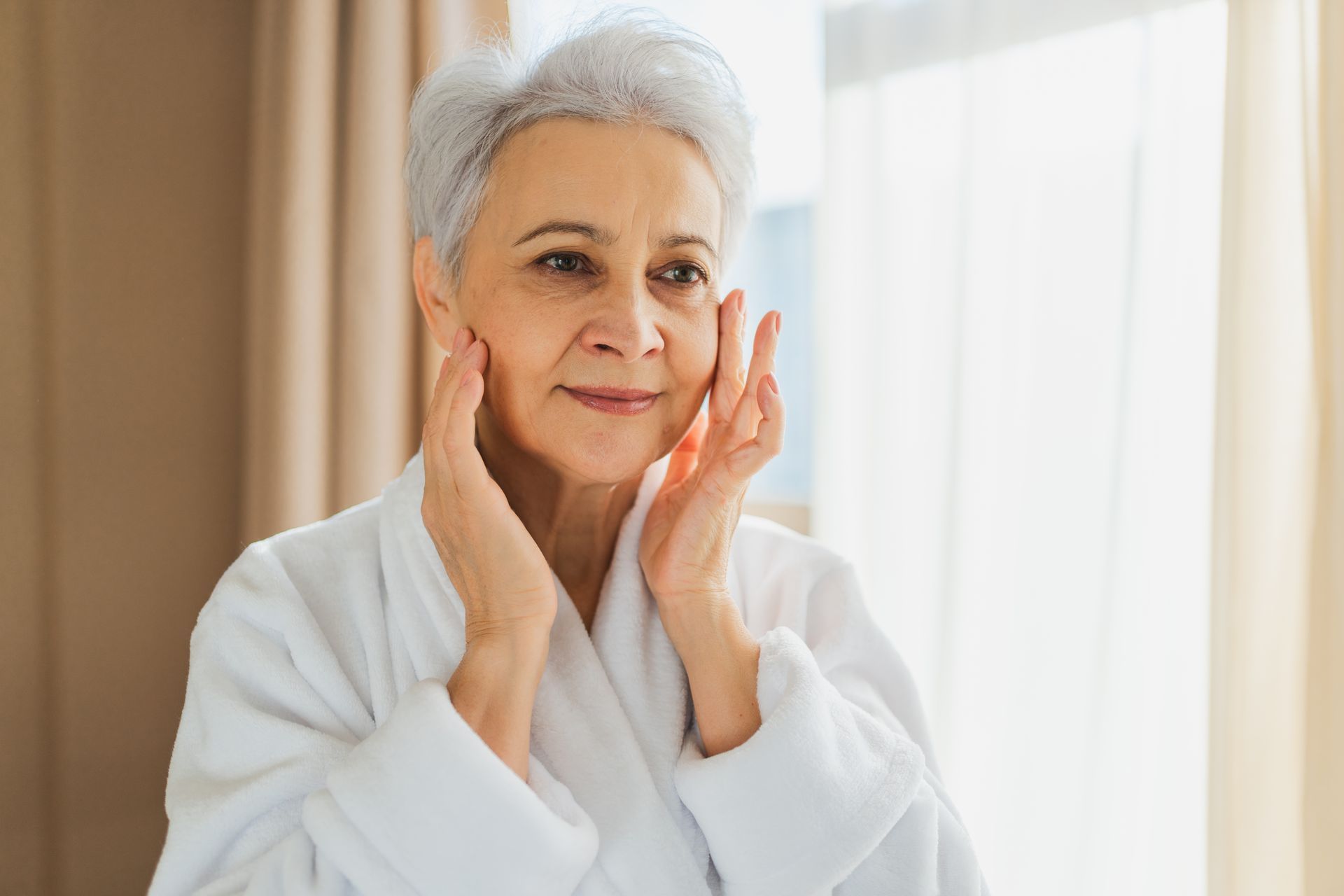 A woman with white hair in a robe gently touches her face near a window.