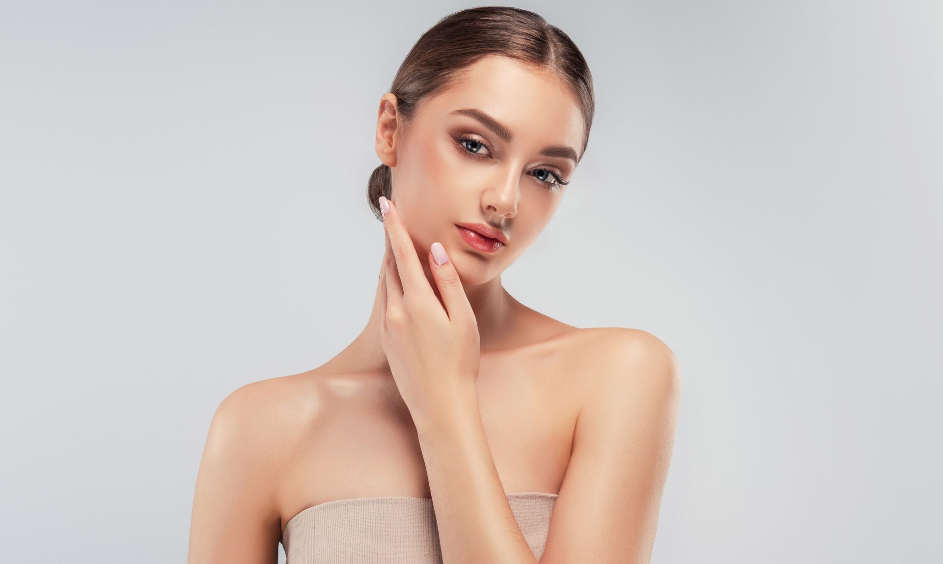 Woman with smooth skin, hand to neck, looking at the viewer with a neutral expression. Neutral background.