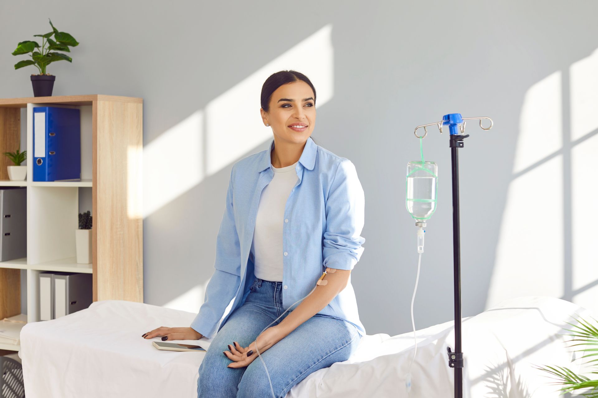 Woman in a clinic, smiling while receiving IV fluids. She's wearing blue, sitting on a bed near a stand.