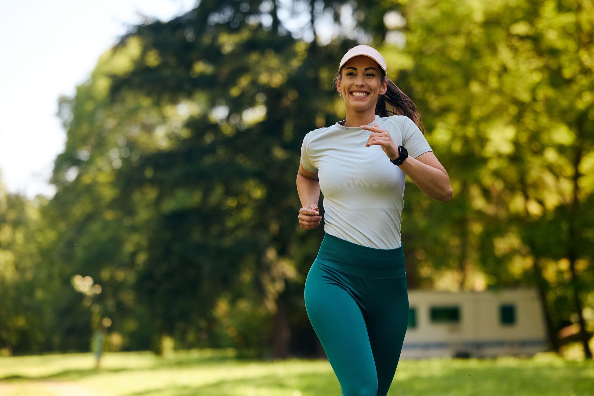 Woman in teal leggings and a cap running in a park, smiling.