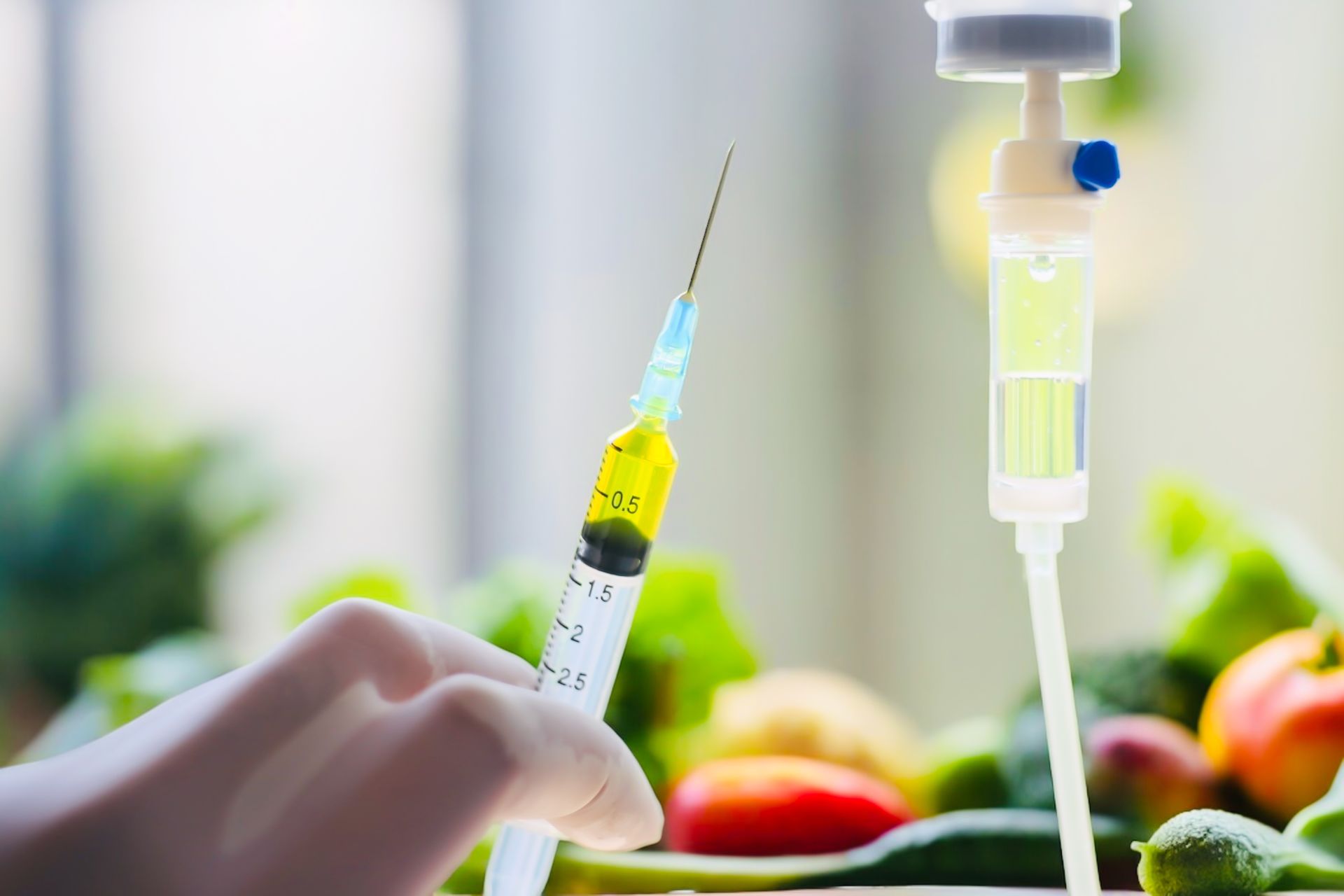 Gloved hand holding a syringe with yellow liquid, next to an IV drip with green fluid; produce in background.