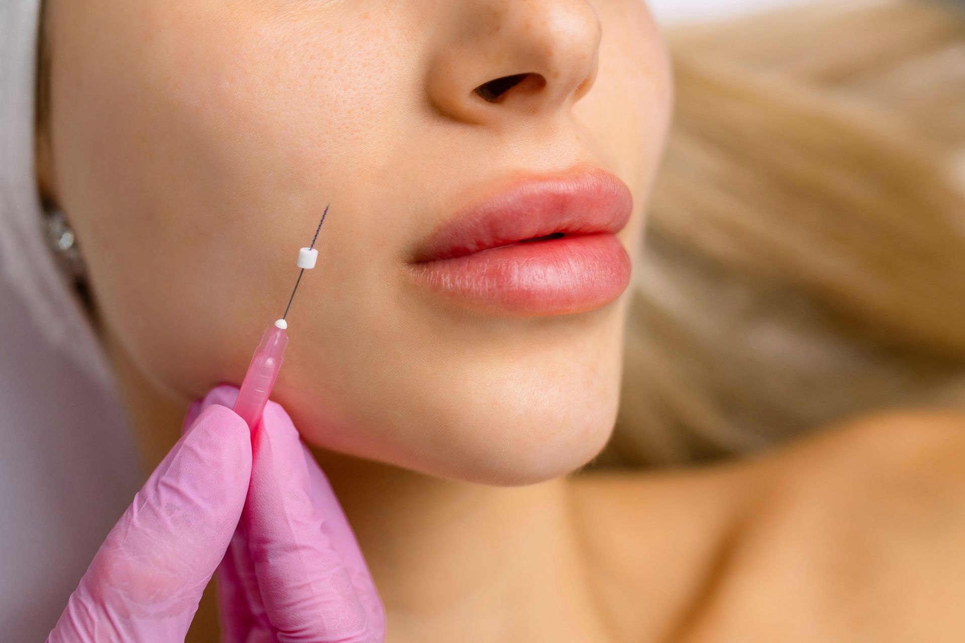 Woman receiving a cosmetic filler injection in her cheek, with pink gloved hand holding the needle.